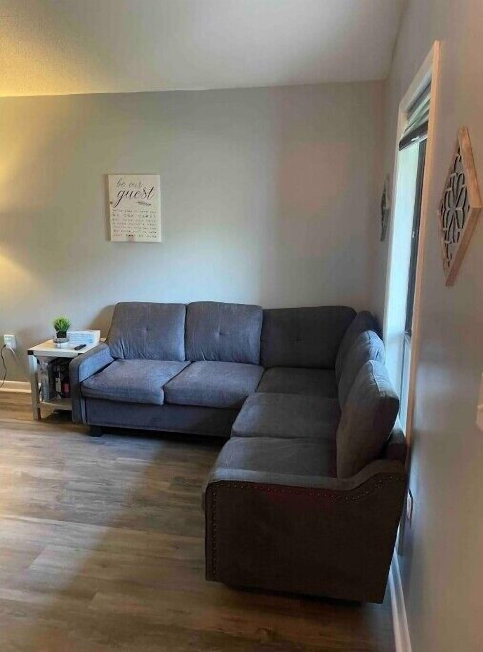Ein Wohnzimmer mit einer grauen Eckcouch, einem kleinen Beistelltisch mit einer Pflanze und einem an der Wand befestigten Schild.
