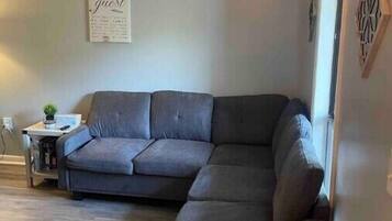 Ein Wohnzimmer mit einer grauen Eckcouch, einem kleinen Beistelltisch mit einer Pflanze und einem an der Wand befestigten Schild.
