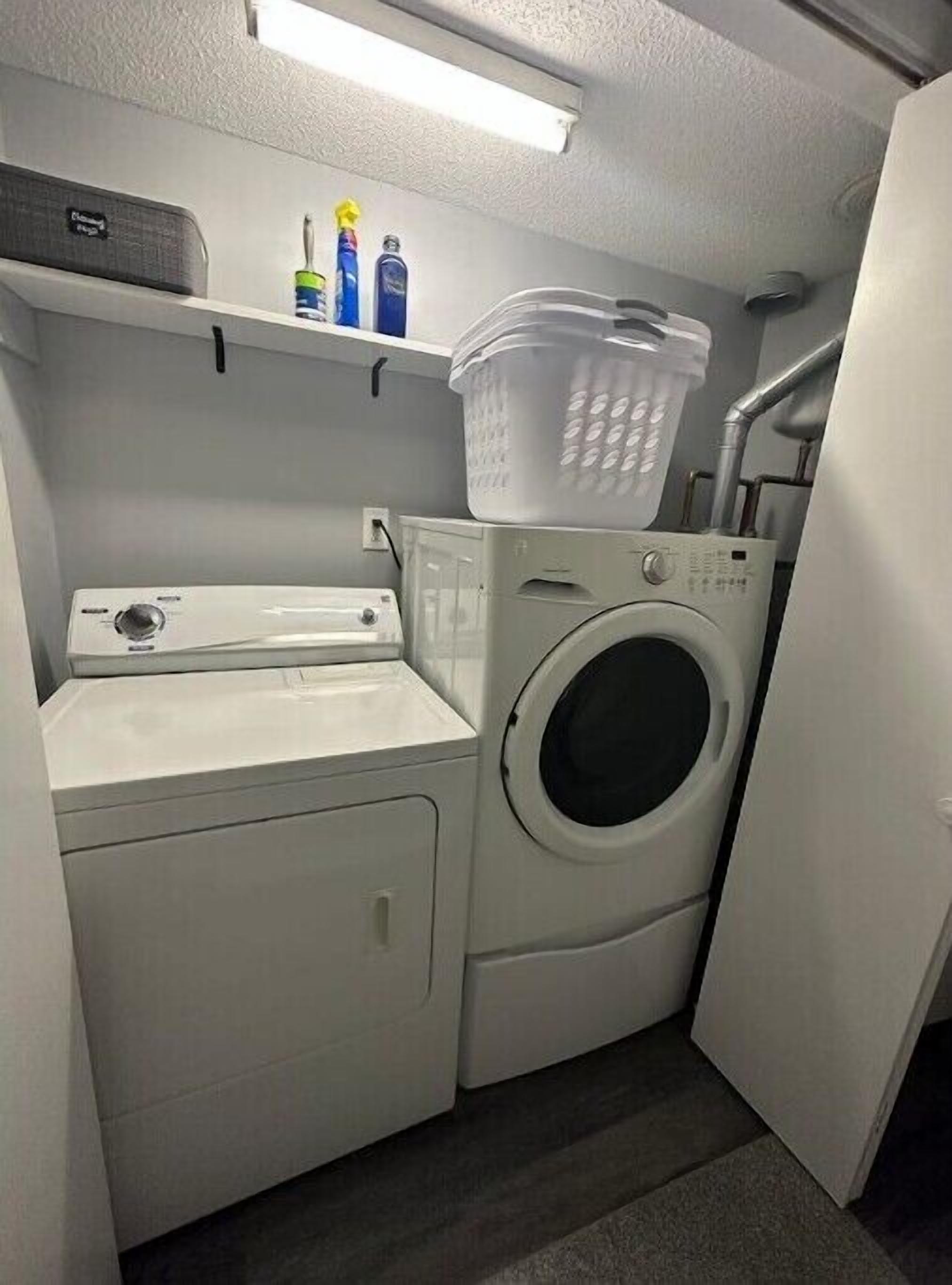 A compact laundry area with a washing machine and dryer, a shelf with cleaning supplies, and a plastic laundry basket.