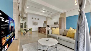 A modern living room with a flat-screen TV, a sofa, a coffee table, and a kitchen area in the background. - Downtown Apt: Pool, Free Breakfast & Valet (Dallas)
