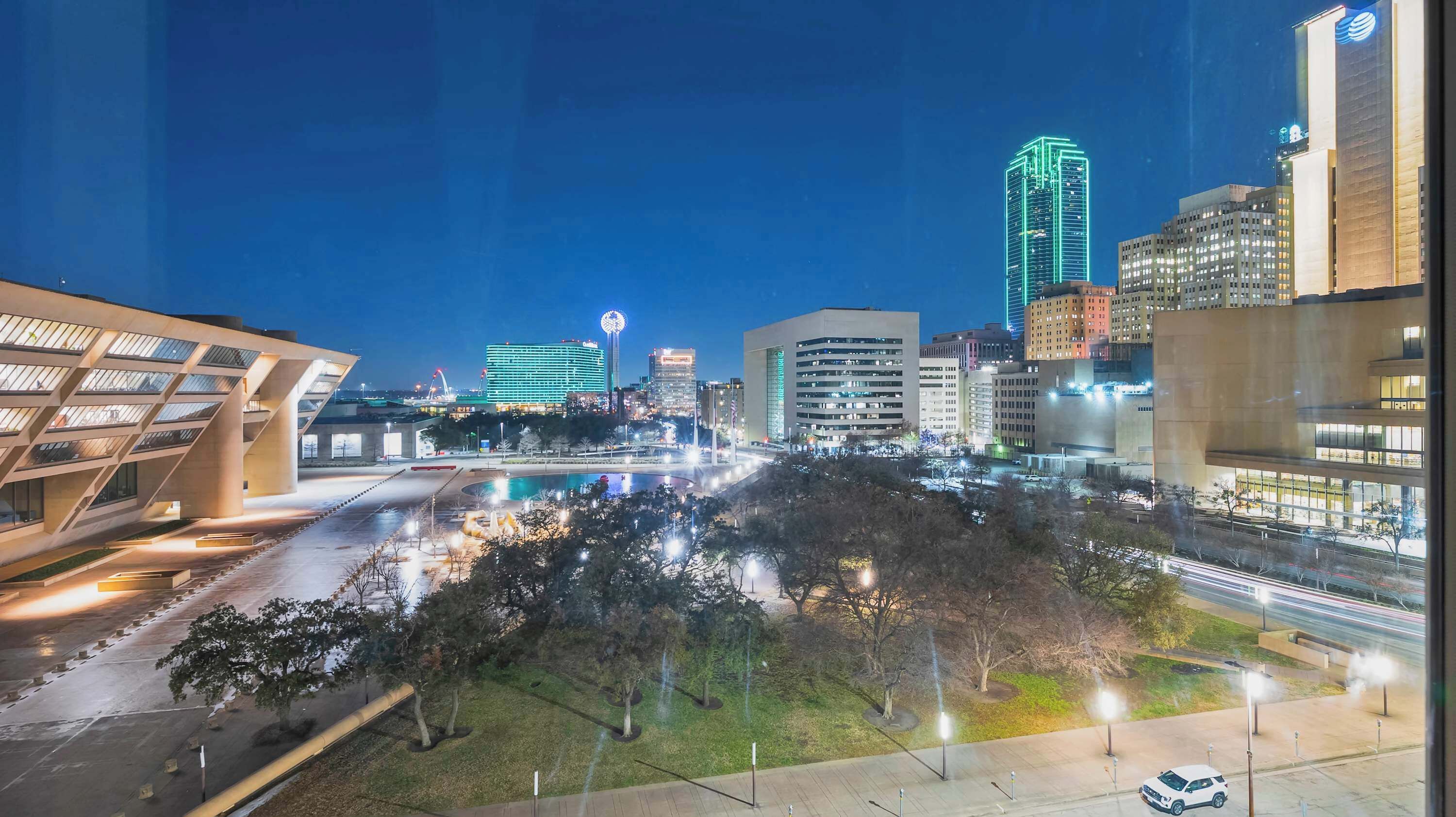 A nighttime cityscape with modern buildings, a large illuminated Ferris wheel, and a well-lit public square.