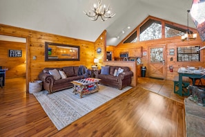 A log cabin interior with a large wooden dining table, two leather sofas, a chandelier, and a kitchen area.