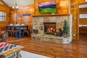 A cozy lodge interior with a stone fireplace, a television mounted above it, and a dining area with a checkered table.