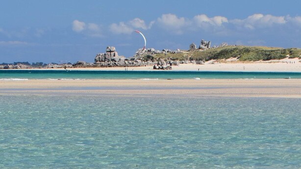 Ein Strand mit klarem türkisblauem Wasser, einem Sandufer und Felsen in der Ferne.