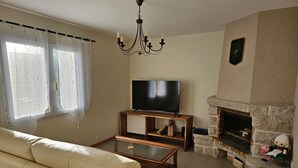 Ein Wohnzimmer mit einem beigefarbenen Sofa, einem Glastisch, einem Flachbildfernseher auf einem Holzboden, einem Kamin aus Stein und einer Kristallleuchter.