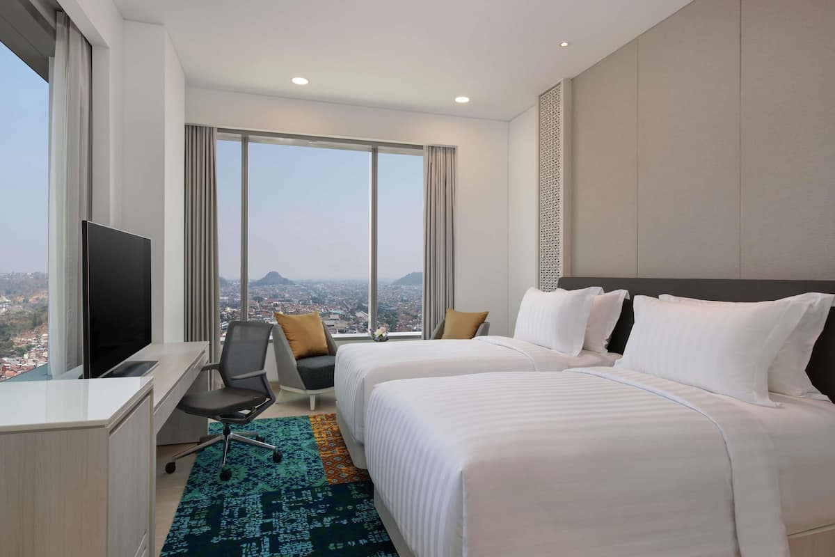 A hotel room with two beds, a desk with a TV, a chair, and large windows offering a city view.