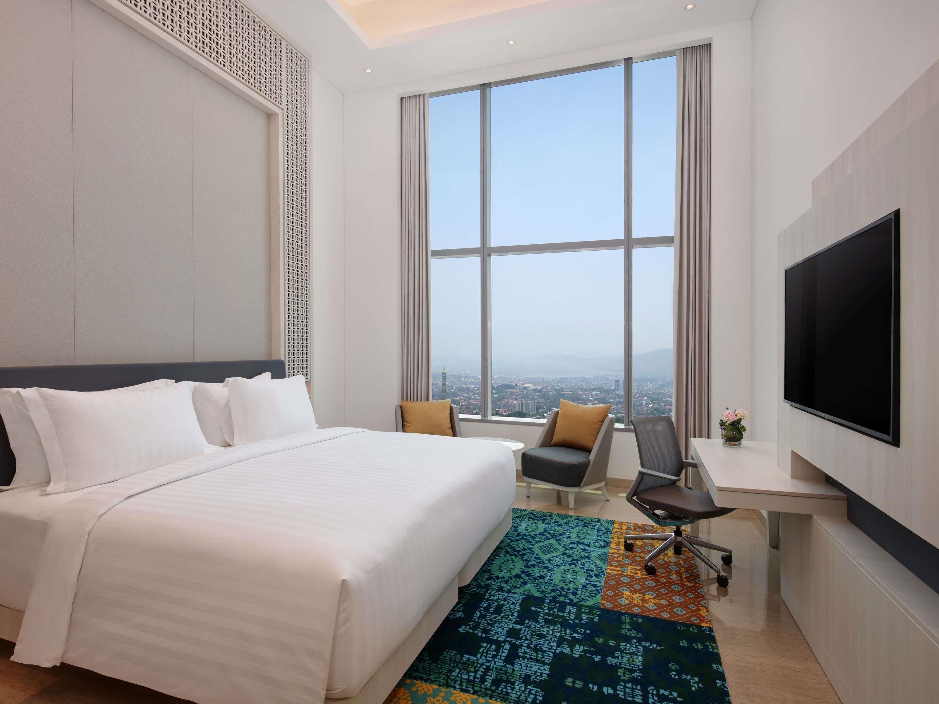 A hotel room with a large bed, a desk, a chair, a TV, and a view of the city.