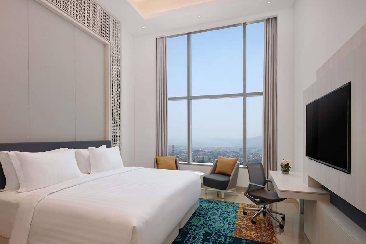 A hotel room with a large bed, a desk, a chair, a TV, and a view of the city.