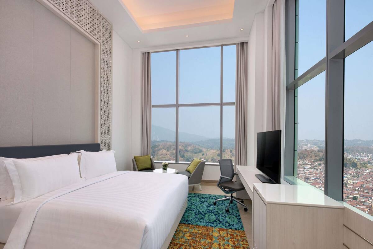A hotel room with a large bed, a desk with a TV, and a view of a cityscape through large windows.