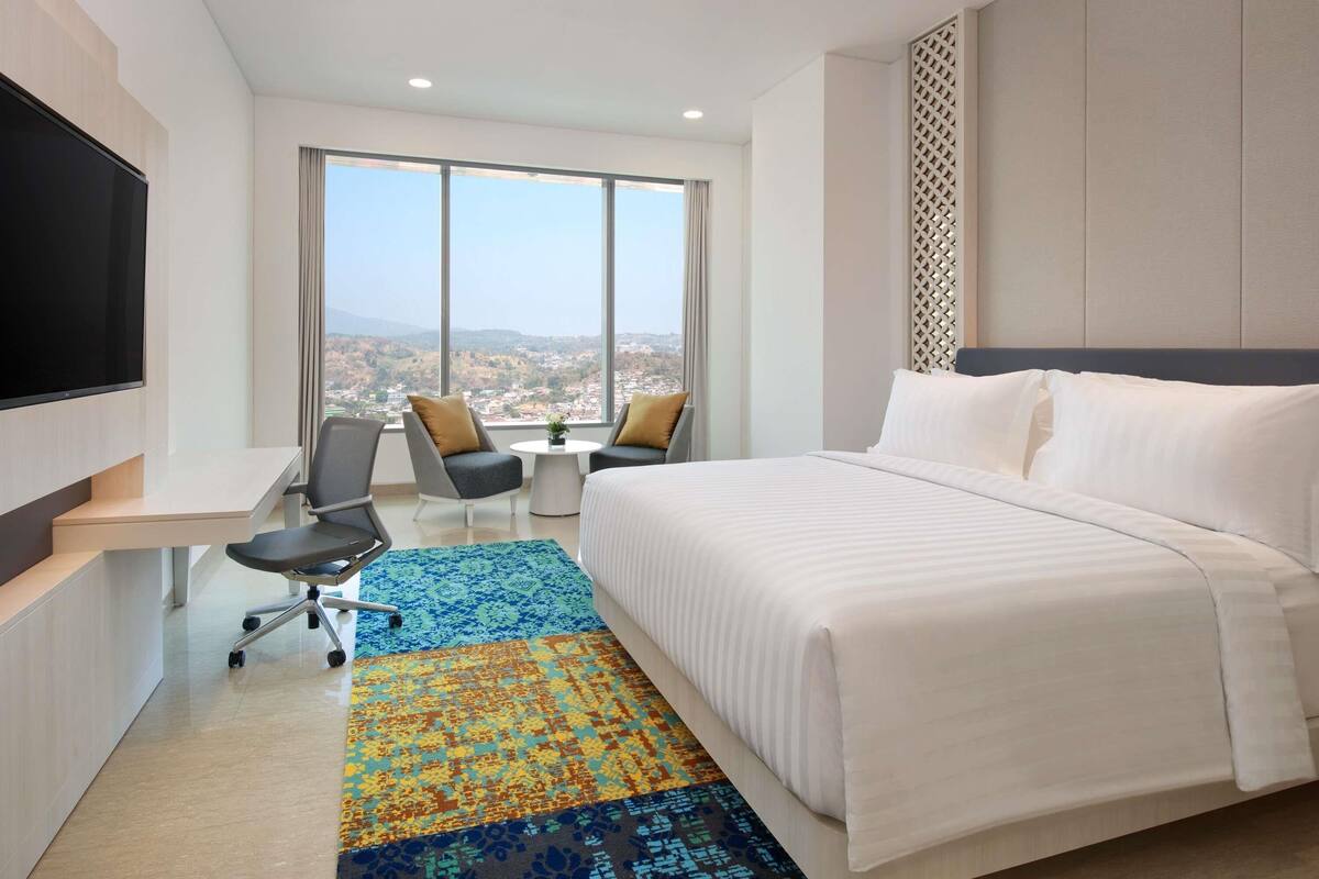 A hotel room with a bed, a desk with a chair, a TV, and a large window offering a city view.