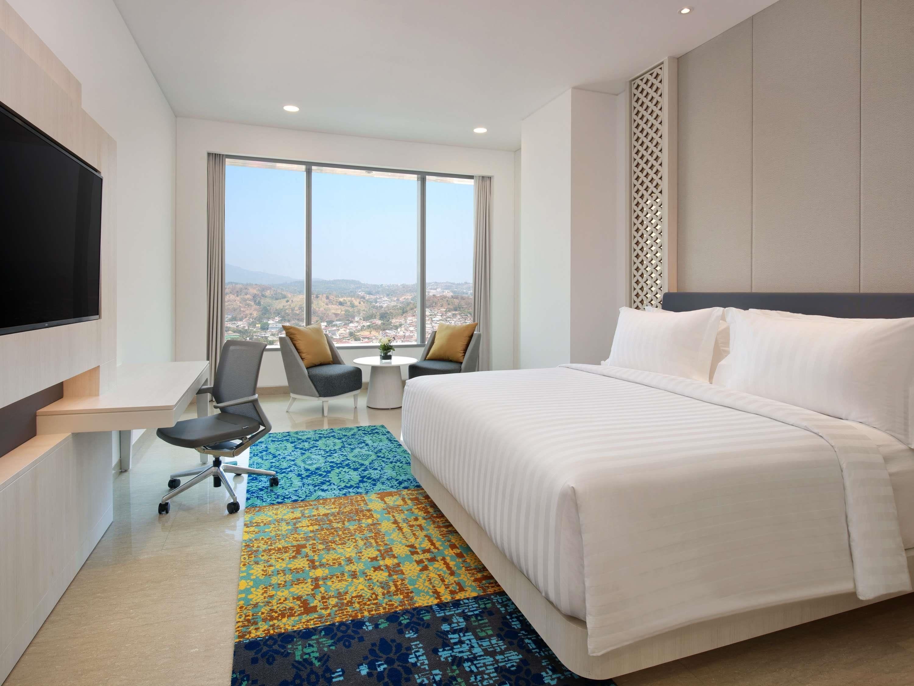 A hotel room with a bed, a desk with a chair, a TV, and a large window offering a city view.