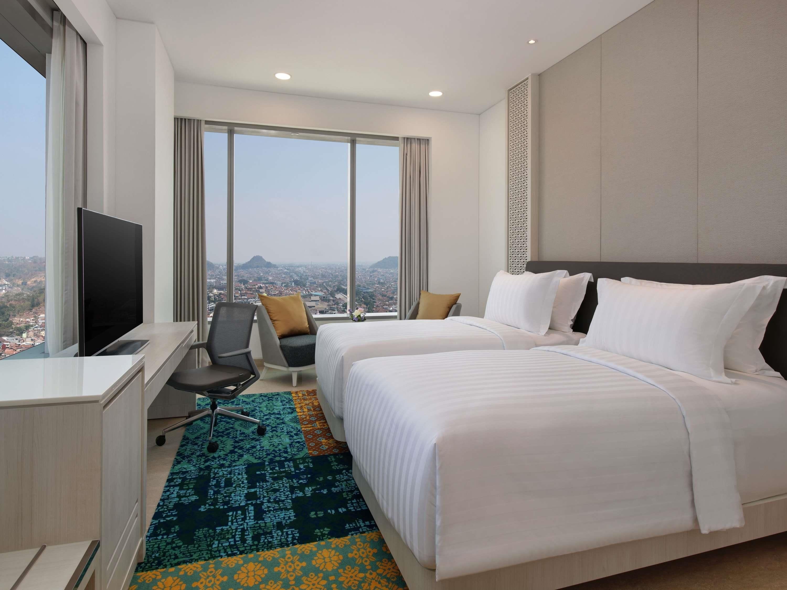Quarto de hotel com duas camas, uma escrivaninha com televisão, uma cadeira e amplas janelas com vista para a cidade.