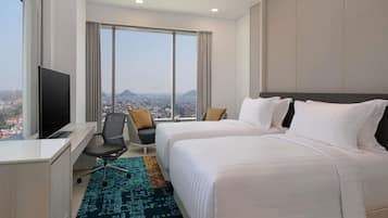 Quarto de hotel com duas camas, uma escrivaninha com televisão, uma cadeira e amplas janelas com vista para a cidade.