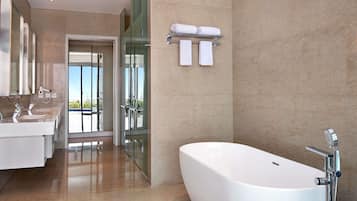 A modern bathroom with a large bathtub, two sinks, a glass shower enclosure, and a view through the glass door.