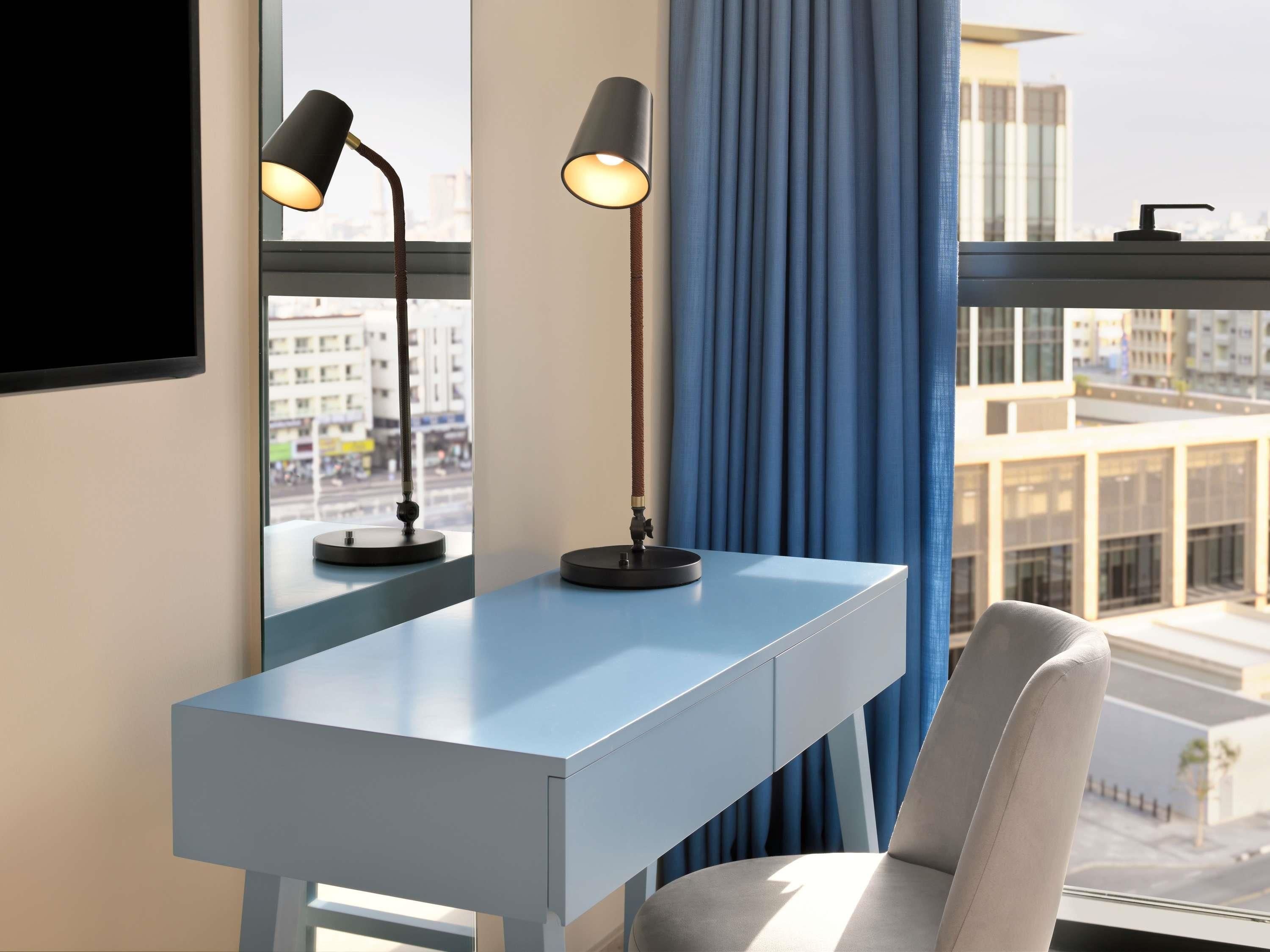 A modern desk with two lamps, a chair, a window with blue curtains, and a city view.