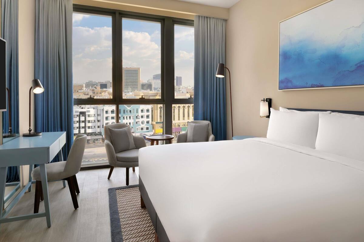 Une chambre d’hôtel avec un grand lit, un bureau, deux chaises, une petite table, une lampe, un tableau et une vue sur la ville grâce à de grandes fenêtres.