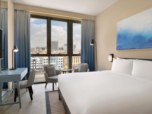 A hotel room with a large bed, a desk, two chairs, a small table, a lamp, a painting, and a view of the city through large windows. - Mercure Dubai Deira (Dubai)