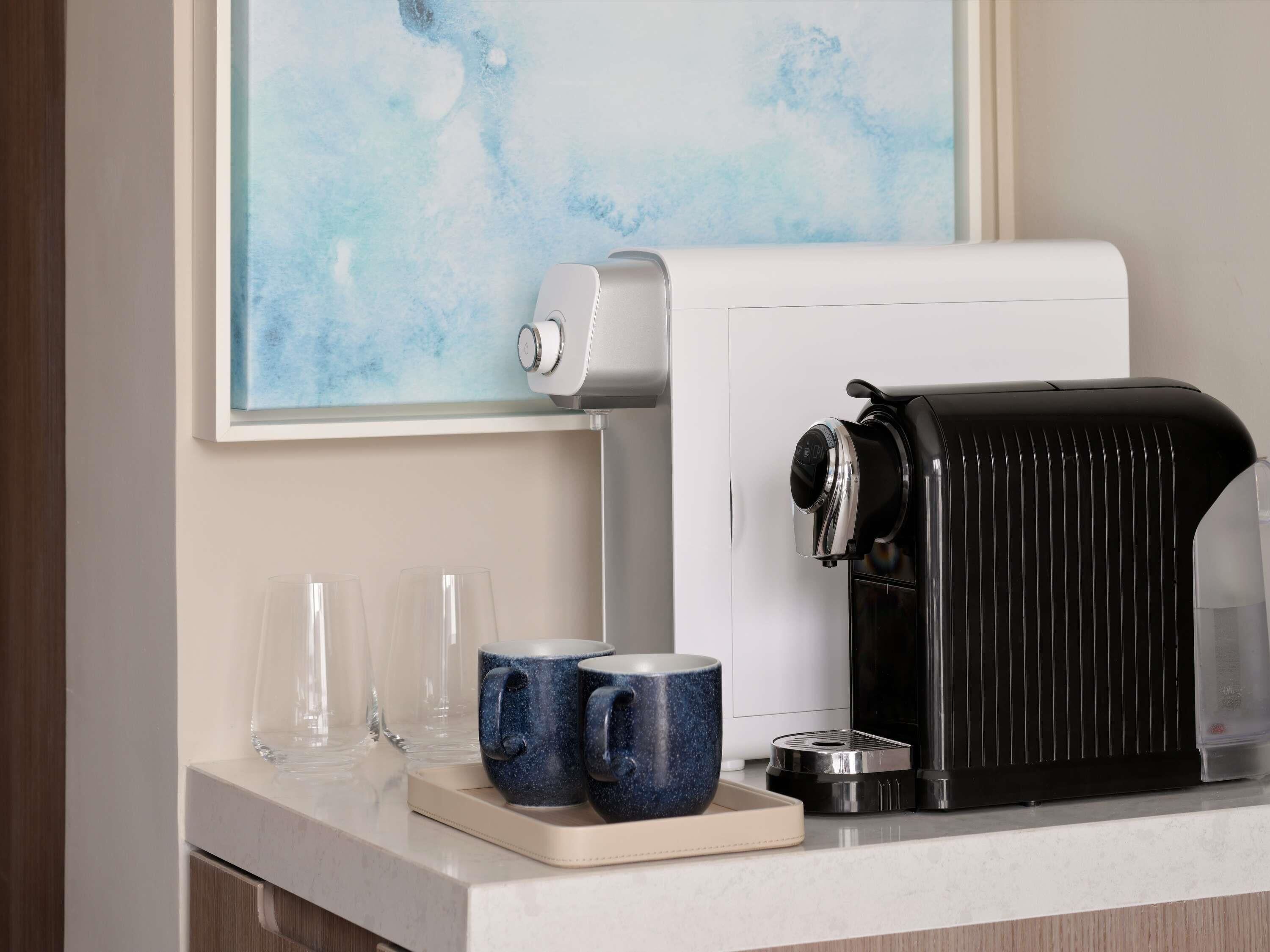 A coffee machine, two blue mugs, and a tray with glasses on a countertop.
