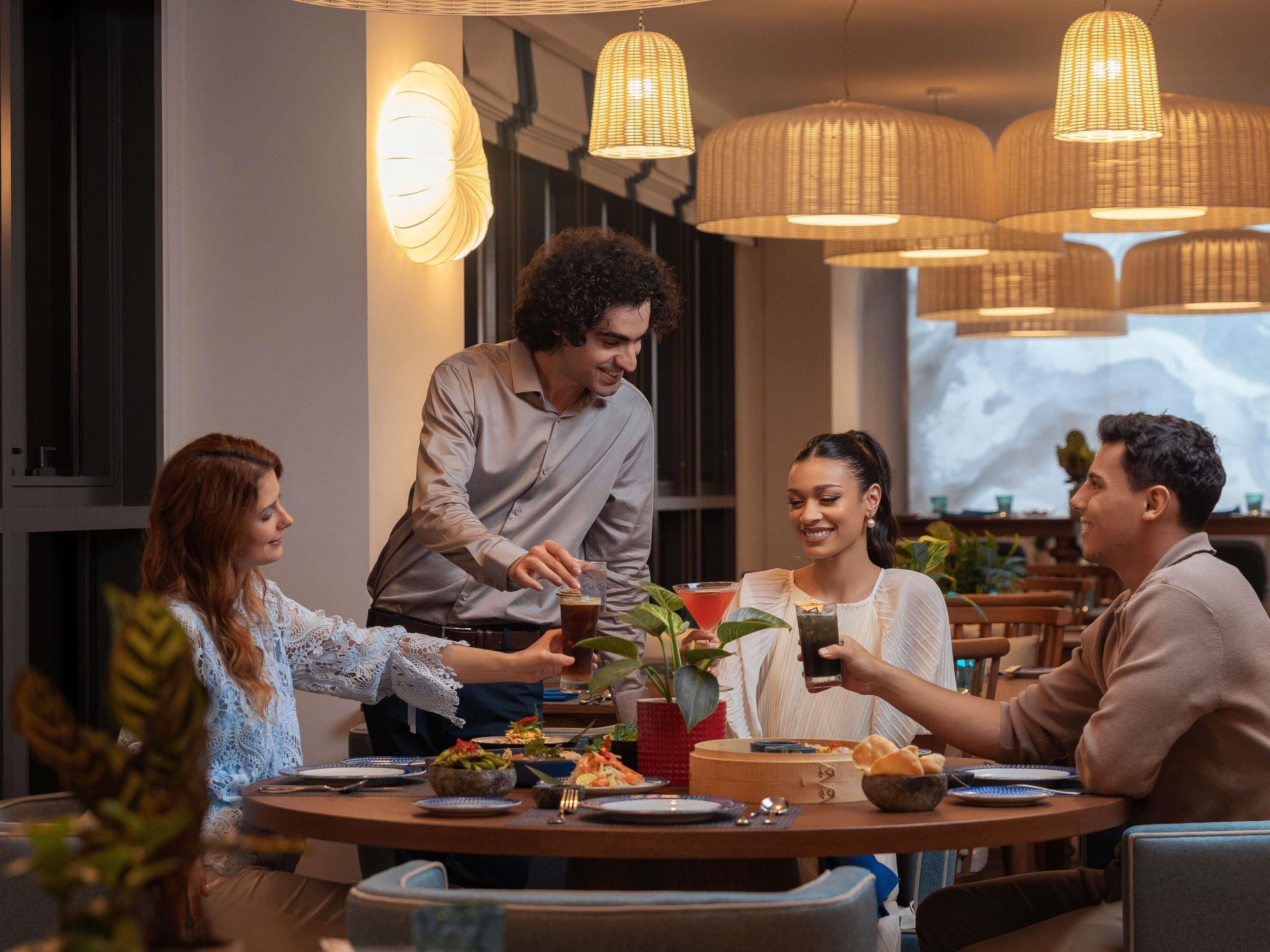 A group of people dining at a restaurant with a modern interior, including pendant lighting and a view of the cityscape.