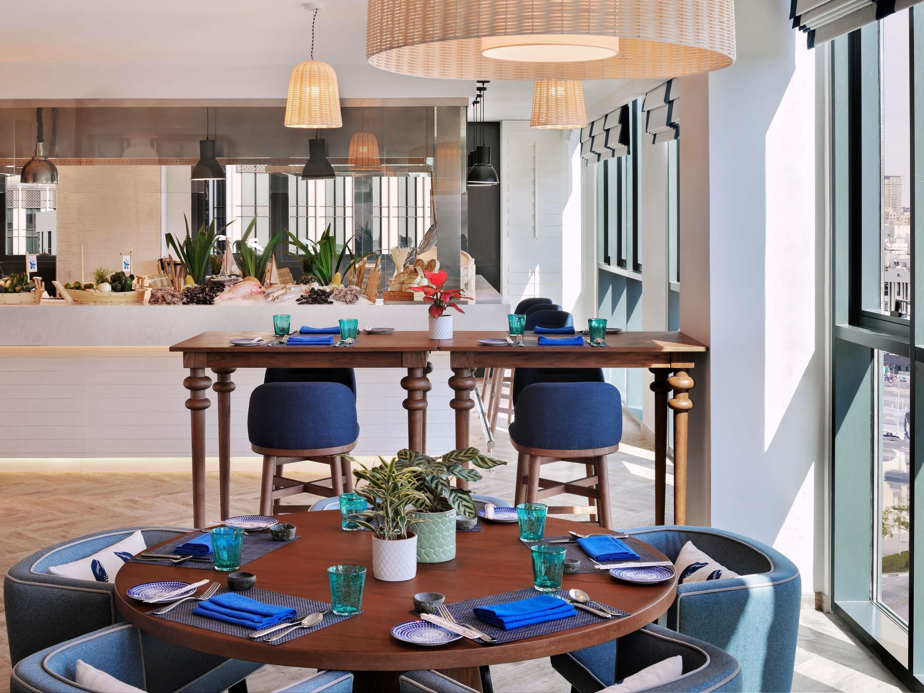 Un comedor moderno con una mesa redonda dispuesta para cuatro personas, sillas tapizadas en azul, una planta en maceta en el centro y una variedad de vajilla.
