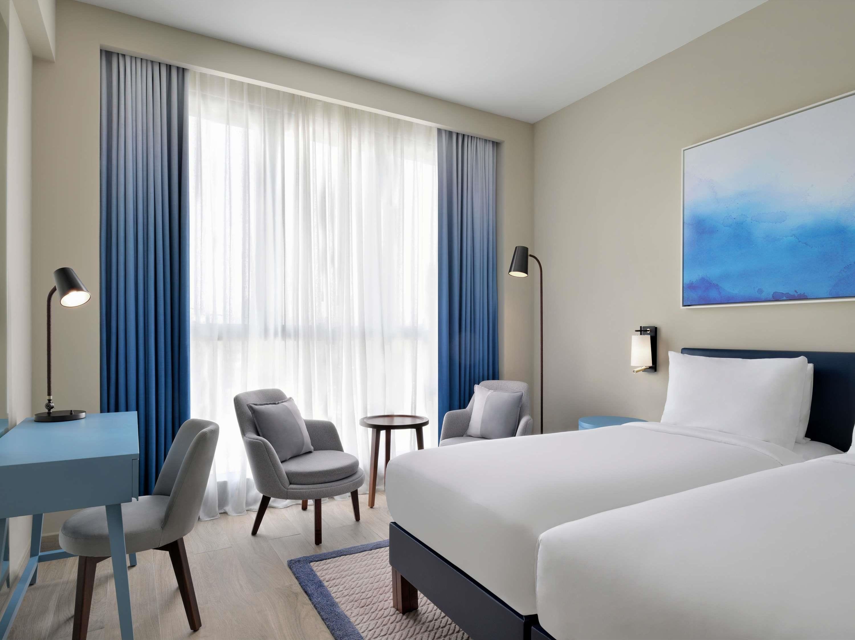 Quarto de hotel com uma cama, duas cadeiras, uma escrivaninha, um abajur e um quadro na parede.