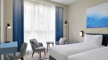 Quarto de hotel com uma cama, duas cadeiras, uma escrivaninha, um abajur e um quadro na parede.