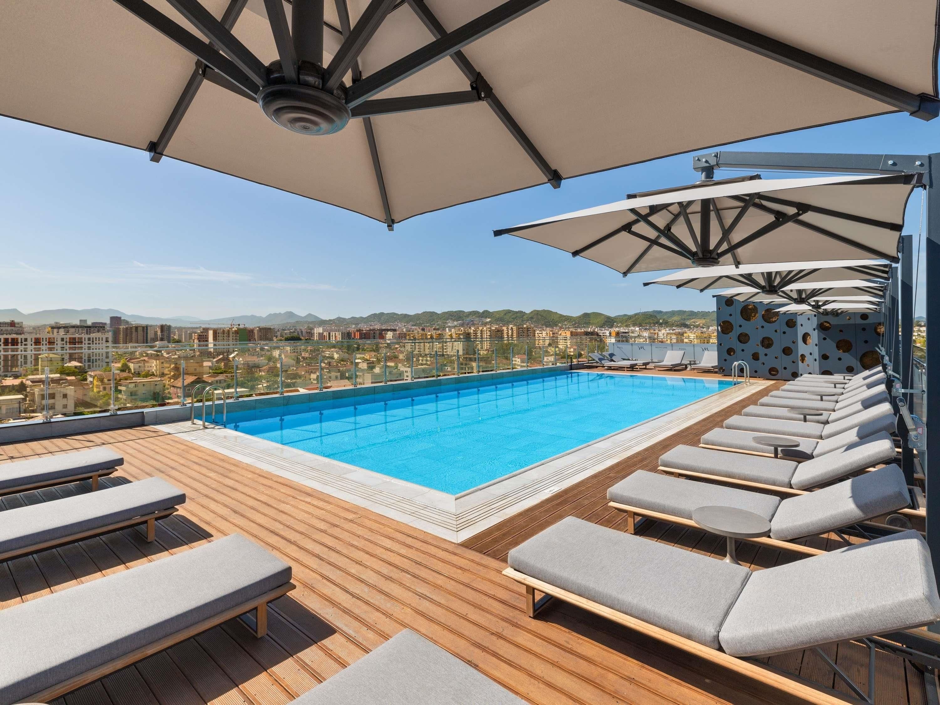 A rooftop pool with lounge chairs and umbrellas, overlooking a cityscape.