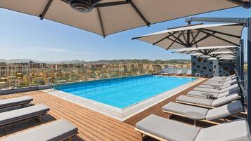 A rooftop pool with lounge chairs and umbrellas, overlooking a cityscape.
