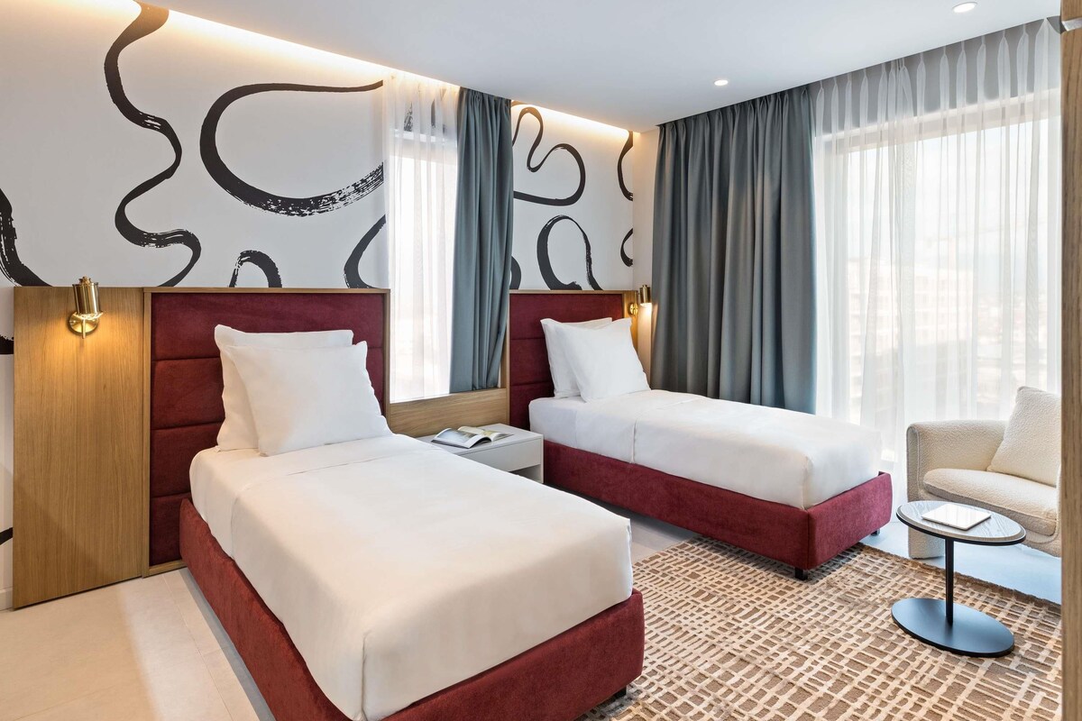 A hotel room with two beds, a small table, a chair, and a large window with curtains.
