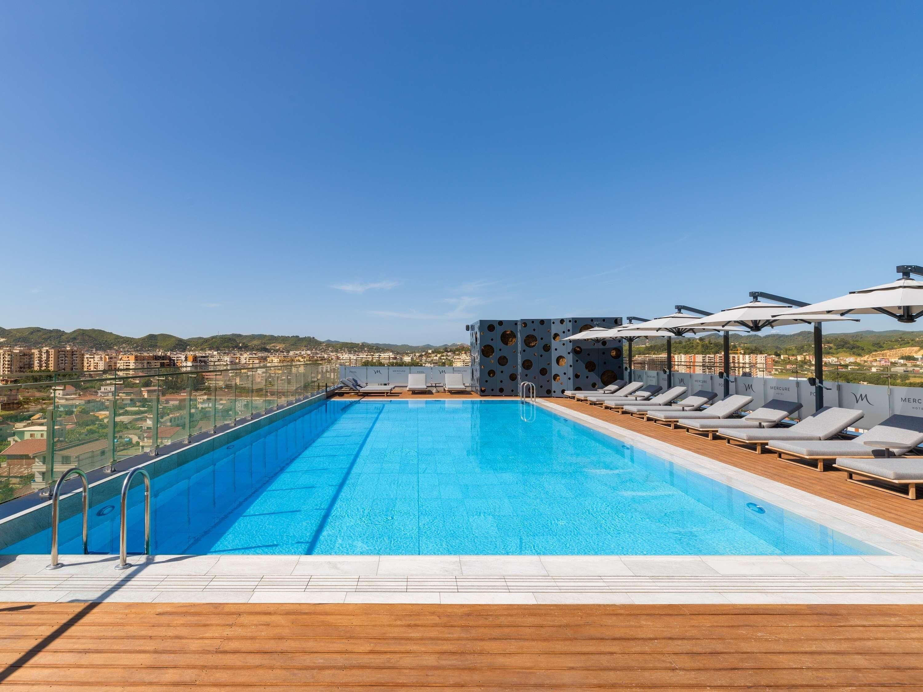 A rooftop swimming pool with lounge chairs and umbrellas, surrounded by a cityscape view.