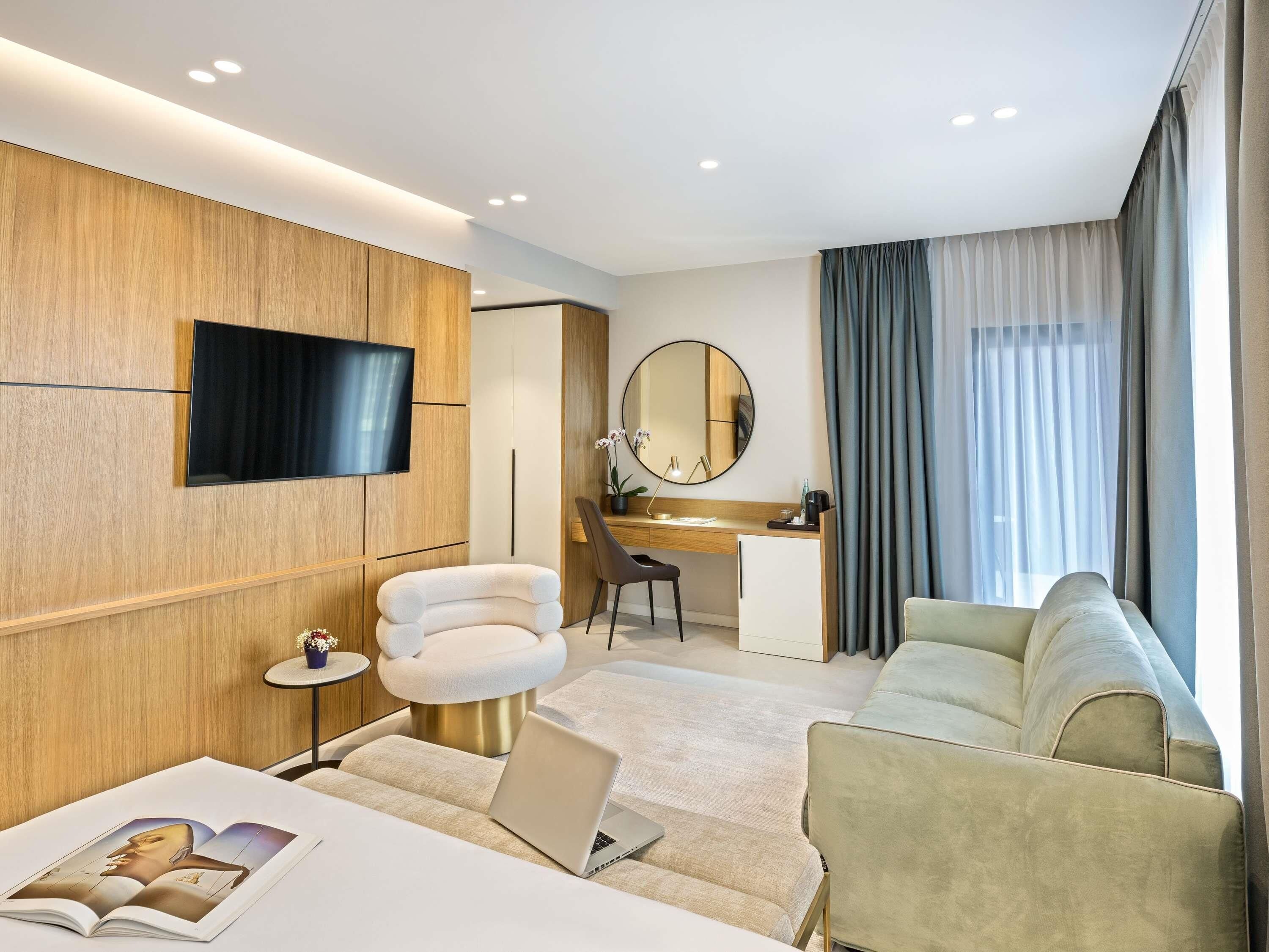 Una habitación de hotel moderna con una televisión de pantalla plana, un espejo redondo, un escritorio con una silla, un sofá y un ordenador portátil sobre una mesita acolchada.