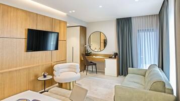Una habitación de hotel moderna con una televisión de pantalla plana, un espejo redondo, un escritorio con una silla, un sofá y un ordenador portátil sobre una mesita acolchada.