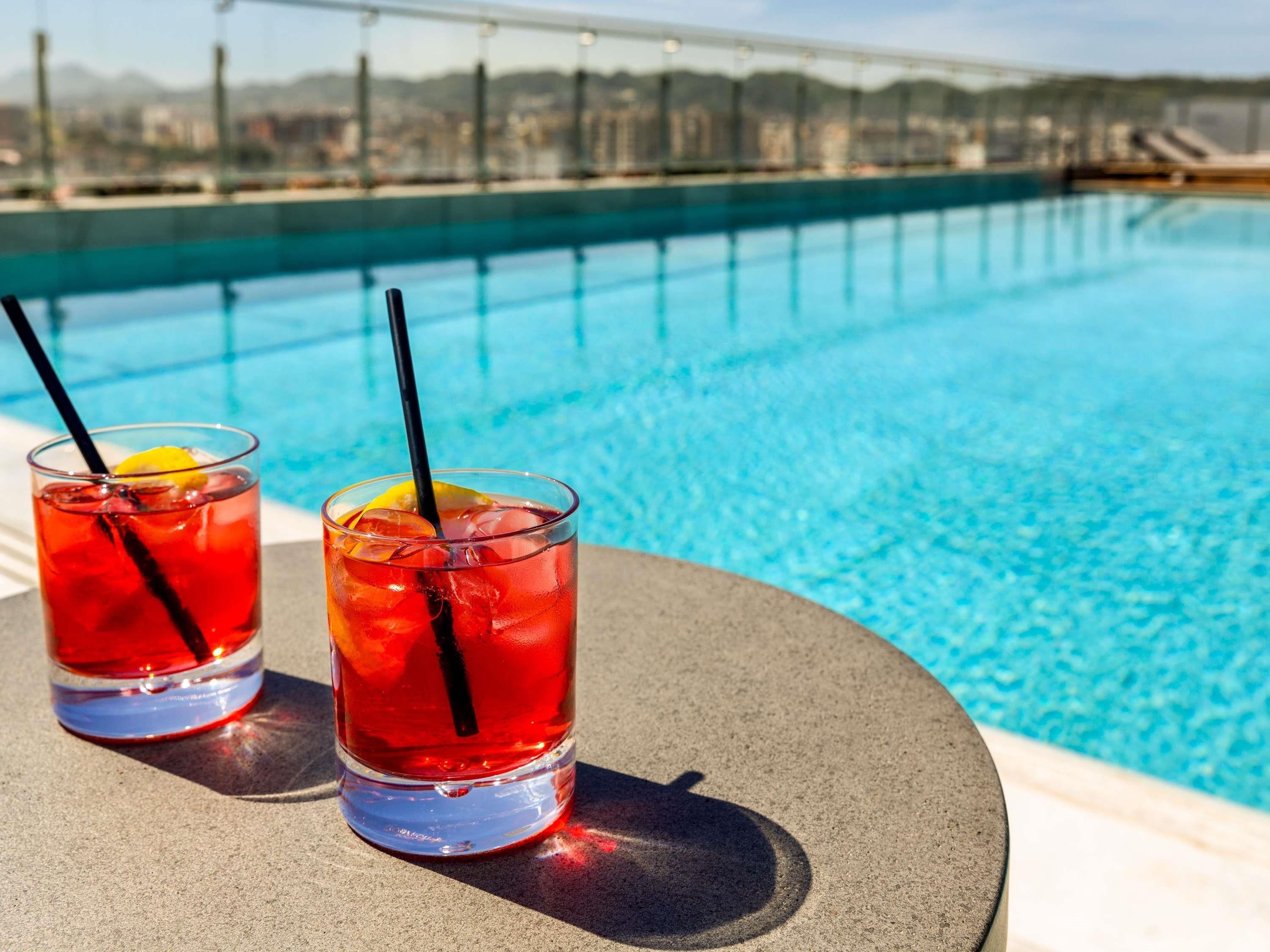 Dos vasos de bebida roja con pajitas y rodajas de limón sobre una mesa redonda junto a una piscina con vistas a la ciudad.