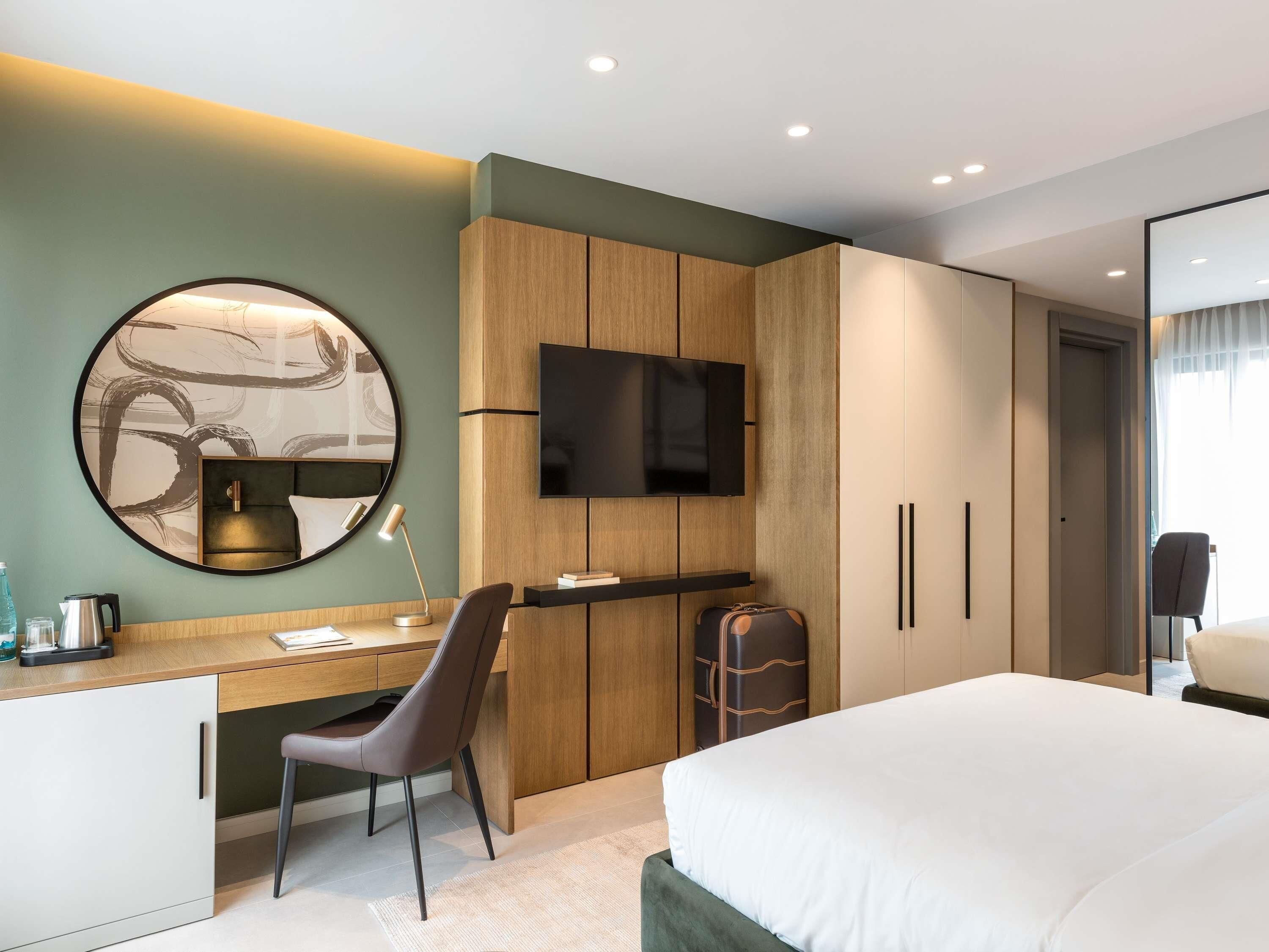 A hotel room with a bed, a desk with a chair, a TV mounted on the wall, and a wardrobe.