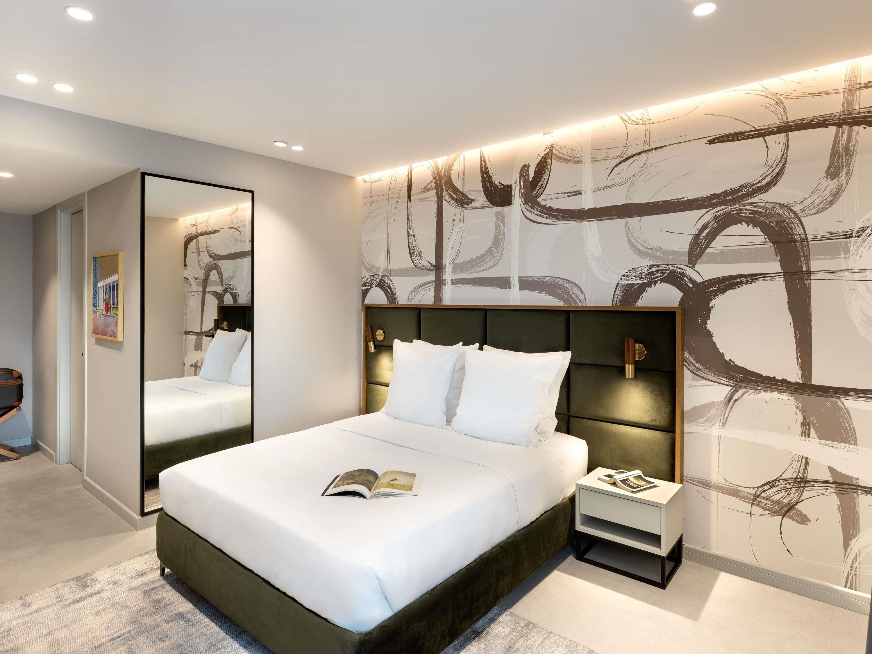 A modern hotel room with a large bed, bedside tables, a mirror, and abstract wall art.
