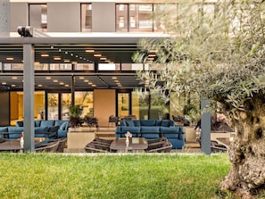 An outdoor seating area with blue sofas and wicker chairs, a large tree, and a modern building with large windows in the background. - Mercure Tirana (Tirana)