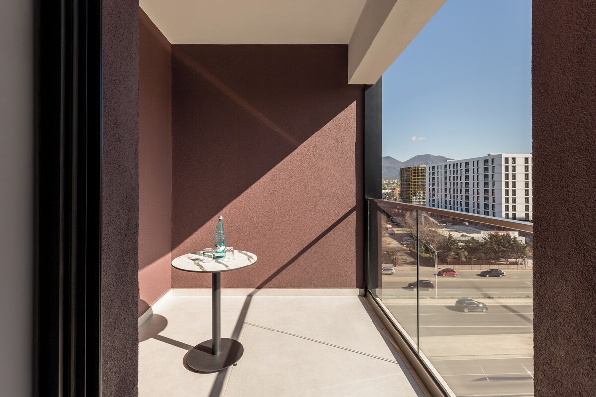 A balcony with a round table and a bottle, overlooking a parking lot and buildings.