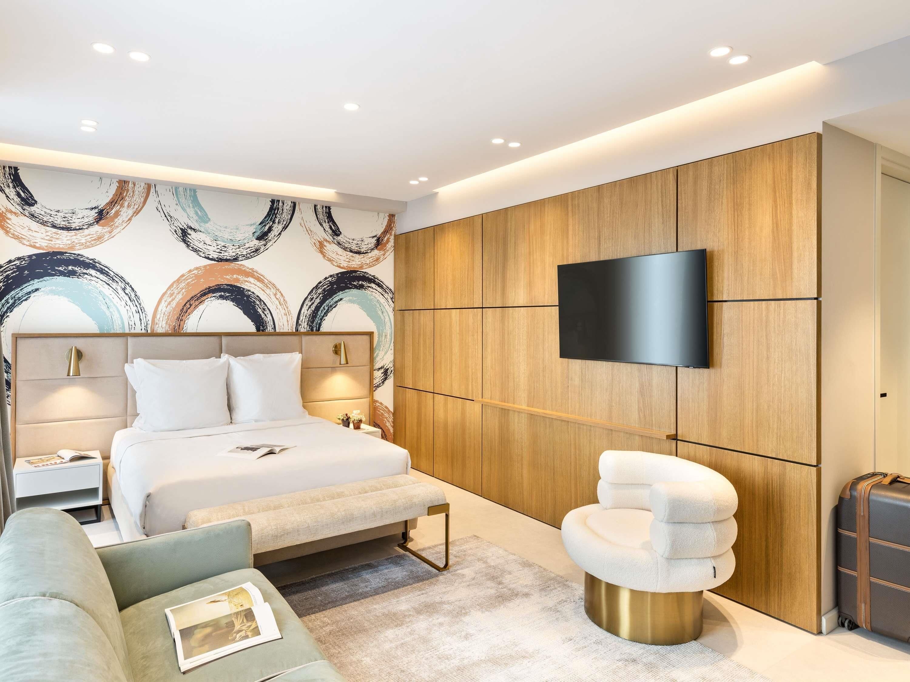 A modern hotel room with a bed, a sofa, a seating area, a television, and a wardrobe.