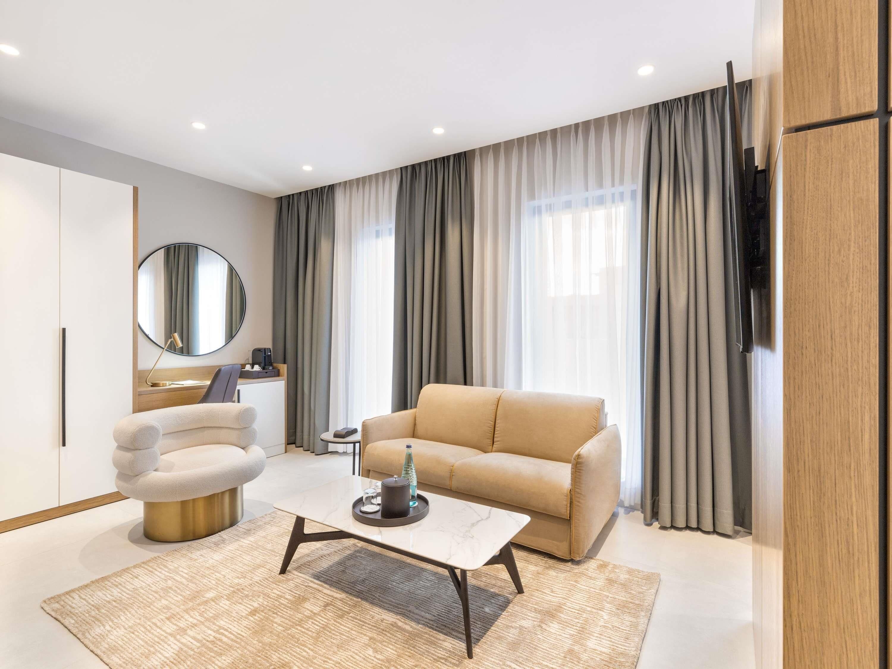 A modern hotel room with a beige sofa, a round armchair, a coffee table, a side table with a phone and a coffee machine, and a large window with curtains.