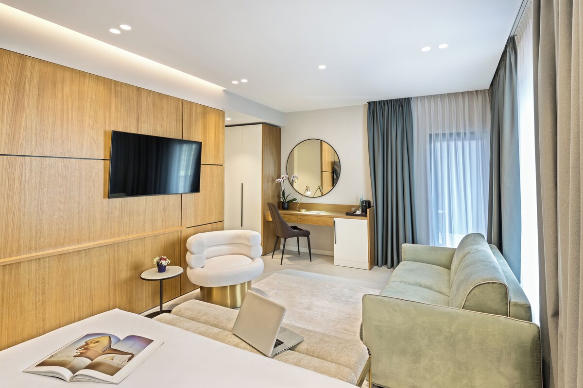 A modern hotel room with a flat-screen TV, a round mirror, a desk, a comfortable sofa, and a laptop on the bed.