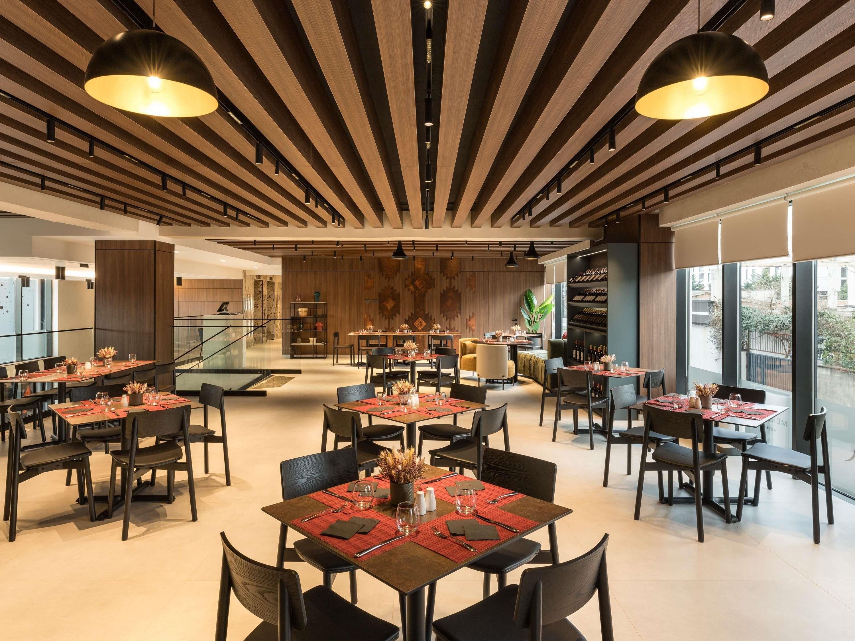 A modern dining area with multiple tables and chairs, wooden ceiling panels, and pendant lighting.