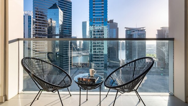 Un balcon avec deux chaises au design épuré, en fil de fer noir, et une petite table ronde sur laquelle se trouvent une tasse et un livre. Une rambarde en verre sépare le balcon de la vue sur la ville en contrebas.