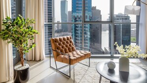 A modern living room with a large window, a brown leather chair, a potted plant, a round table with a vase, and a lamp. - Silkhaus Stunning Prime Design 1 BDR Gym Pool (Dubai)