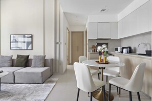 Una sala moderna con un sofá gris, una mesa de comedor redonda con sillas blancas, y una cocina con armarios y electrodomésticos blancos.