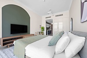 Een moderne hotelkamer met een groot bed, een flatscreen televisie, een houten tv-meubel, een klein tafeltje met een plant, en een keukenruimte zichtbaar door een geopende deur.