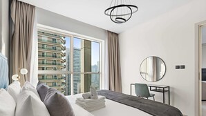 A hotel room with a large bed, a desk, a chair, a round mirror, and a view of high-rise buildings. - Silkhaus Chic in West Avenue Pool Gym Access (Dubai)
