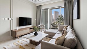 A modern living room with a flat-screen TV, a wooden console, a marble coffee table, a beige sofa, and a view of a cityscape through large windows. - Silkhaus Luxurious New Tower With Pool Gym (Dubai)