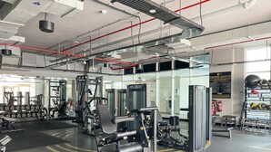 A modern gym with various exercise machines, a mirrored wall, and a well-lit space with visible ceiling pipes and ventilation. - Silkhaus Luxurious Studio in Studio One Tower (Dubai)