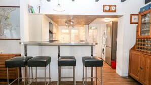 A kitchen with a central island, bar stools, a washing machine, and a wooden cabinet with glassware. - The Cottage (Pwllheli)