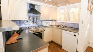 A modern kitchen with stainless steel appliances, wooden cabinets, and a window with bars. - The Cottage (Pwllheli)